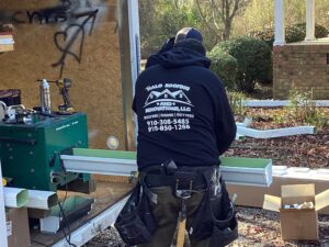 A person in a black hoodie with the logo "Halo Roofing and Renovations, LLC" is working on a gutter installation. They are using a green machine to shape the gutter. Various tools and supplies are scattered around, with a partially constructed building in the background, showcasing expert roofing work.
