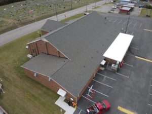 Aerial view of a large brick building with a gray roofing and an adjacent parking lot. A red truck is parked near the entrance, which has a white canopy. A cemetery with aligned gravestones is visible in the background.