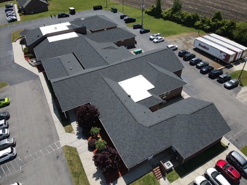 Aerial view of a large building with a dark grey roofing, surrounded by a parking lot filled with cars. The building has several wings and a few trees along its perimeter. There is a Sodexo truck parked nearby on the right side of the image.