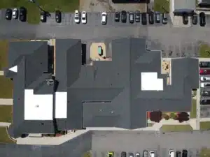 Aerial view of a large building with a gray roofing and several protrusions. Cars are parked in multiple parking lots around the building. There is a small playground area with a sandbox in one corner of the building. The surrounding area includes grassy and paved sections.