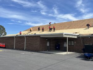 Several workers in red shirts and black pants are installing new roofing material on a large brick building. The roof deck is exposed, and the workers are spread out across the roof, under a clear blue sky. Orange traffic cones are seen to the left.