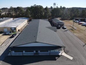 Aerial view of a large commercial building with grey roofing, featuring a black roof and surrounded by a paved lot. Adjacent to the main building, there is a smaller building on the right and a water tower in the background. Several vehicles are parked around the area. Trees are visible beyond the lot.