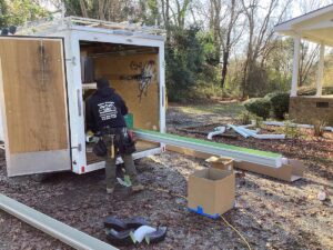 A person stands near an open utility trailer, gathering supplies for a home improvement project. Various materials, including long metal pieces and boxes, are spread around on the ground. The setting is an outdoor area with trees and a house nearby, possibly preparing for some roofing work.