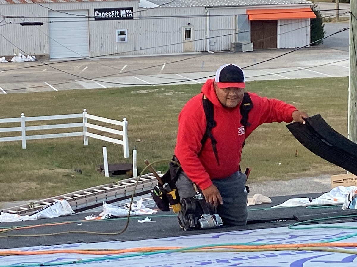 A worker in a red hoodie is kneeling on a roof, holding a piece of Halo Roofing material. He has tools attached to his belt and a nail gun placed beside him. The background shows an empty parking lot, a white fence, and buildings with metal siding.