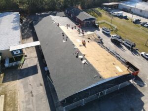 Aerial view of several workers from Halo Roofing repairing the roof of a large building. Part of the roof is stripped down to the wood sheathing, while other sections still have shingles. Nearby, multiple parked vehicles and surrounding buildings are visible.