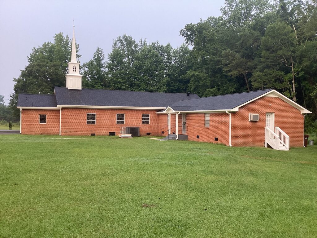 A small brick church with a white steeple and black roofing sits amidst lush greenery. There are a few windows on the sides and an air-conditioning unit on the side wall. The entrance features a small stairway leading up to a white door. The surrounding area is grassy and well-kept.