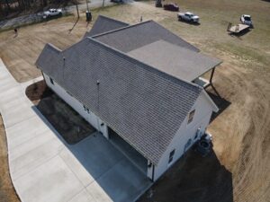 Aerial view of a large single-story house with a brown roofing and white siding. The house sits on an angular, L-shaped foundation and is surrounded by dirt and newly poured concrete paths. Multiple vehicles and people are seen in the background.