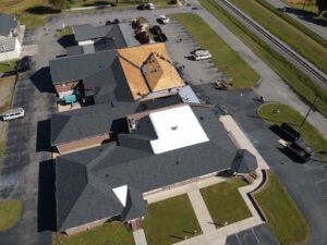 Aerial view of a large, multi-winged building with black and white roof sections, surrounded by a paved parking lot. Multiple vehicles are parked around the building, and there is a road and railway track nearby. Roofing work is visible on the roof amidst ongoing construction.