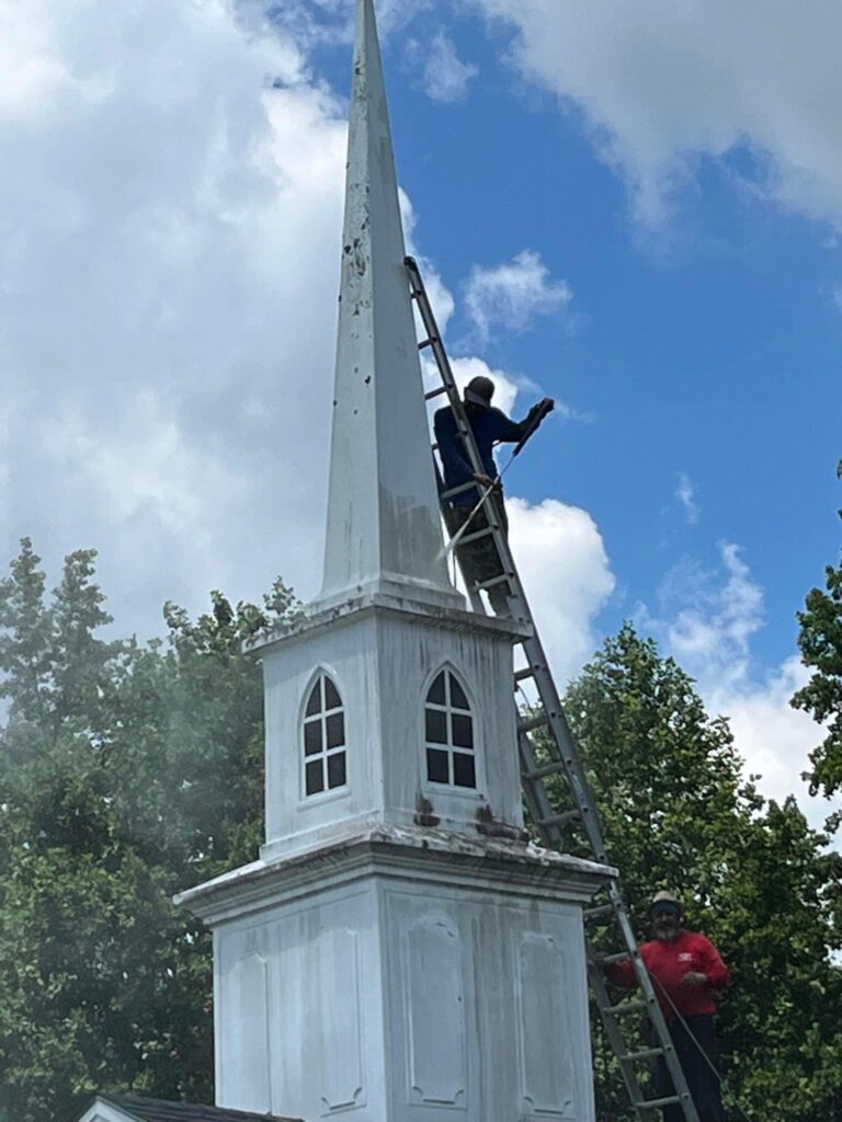 Two workers from Halo Roofing are on ladders cleaning a tall, white church steeple. One person is near the top scrubbing while the other is lower down holding the ladder. The sky is partly cloudy, and trees can be seen in the background.