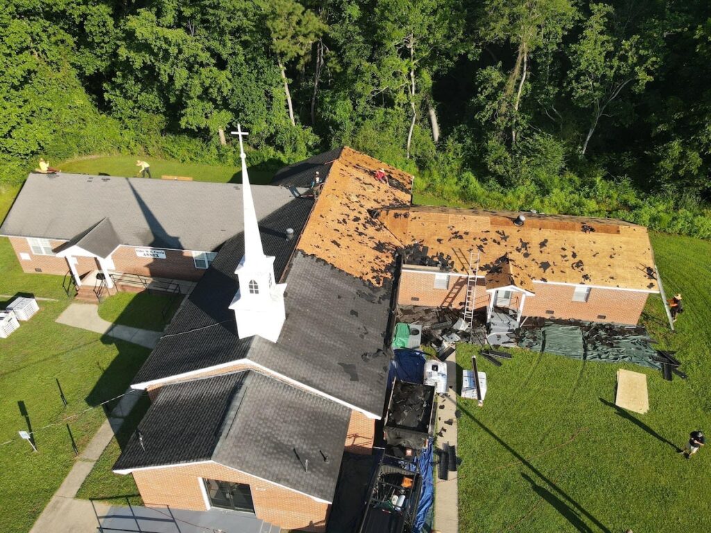 Aerial view of a brick church with a white steeple undergoing roof repairs. Several sections are stripped to the underlayment, and roofing materials are scattered around. Workers from Halo Roofing are seen on the roof and ground, surrounded by green grass and dense trees.