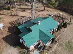 Aerial view of a house with distinctive green roofing in a rural setting. The home is surrounded by a yard with trees, shrubs, and garden structures. A large tree stands near the house, and a pergola or trellis is visible in the yard.