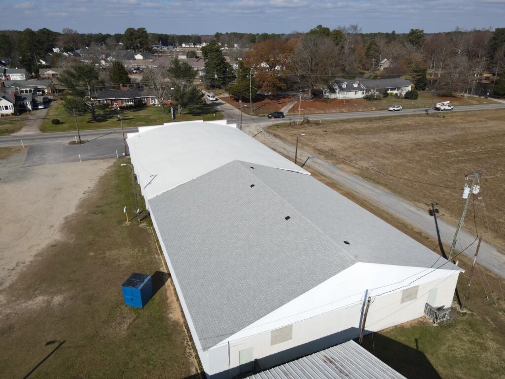 Aerial view of a large, rectangular, single-story building with a light gray roof set in a semi-rural area. Known for its quality roofing, the surroundings include a few scattered houses, vacant lots, and a small patch of woods. The sky is partly cloudy.