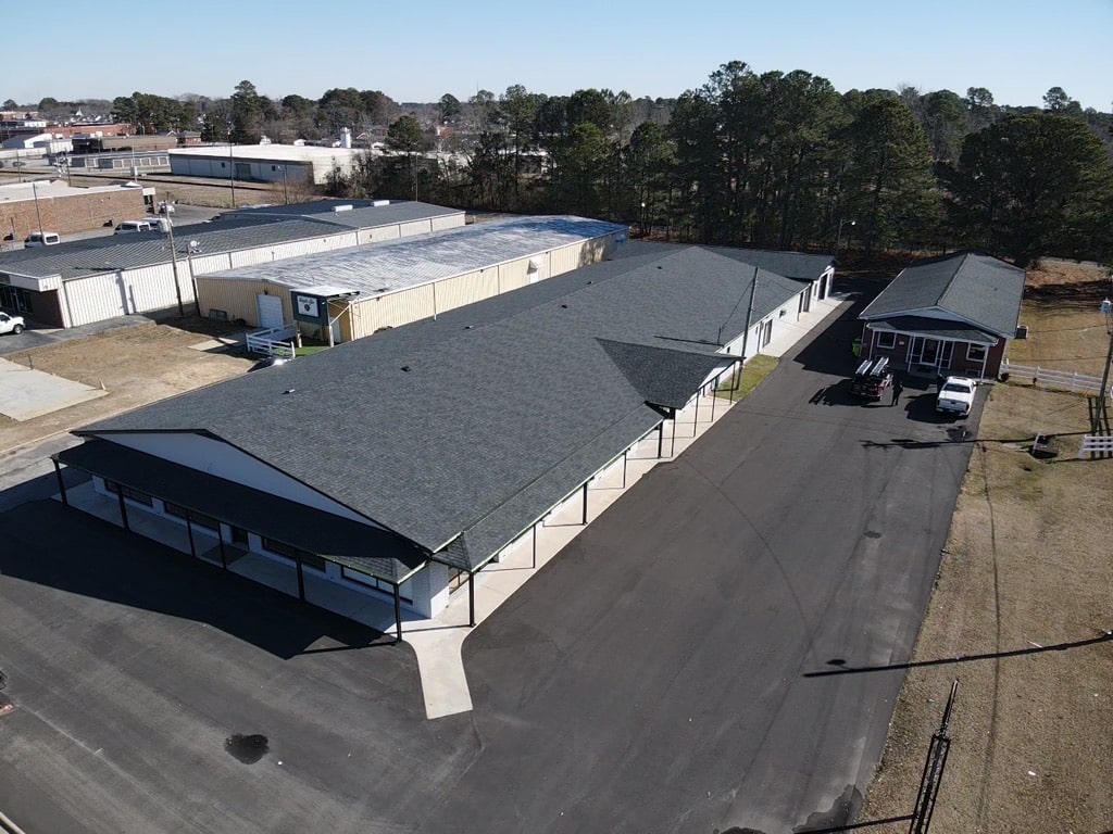 Aerial view of a long, single-story commercial building with black roofing and a white exterior, surrounded by a large paved parking area. There are trees and additional buildings in the background. Adjacent to the main building is a smaller structure with a similar design.