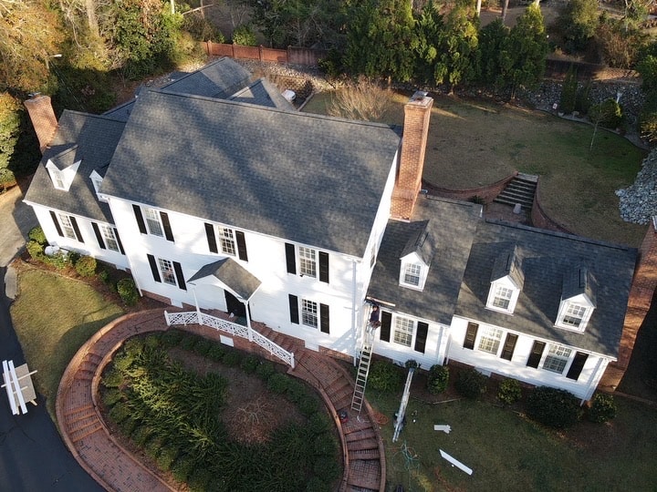 A large white two-story house with black shutters and a gray roofing viewed from above. The house has two chimneys and a surrounding garden with shrubs and a curved brick walkway leading to the entrance. A ladder is propped up against the right side of the house.