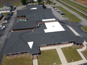 Aerial view of a large building with a complex, multi-wing layout, featuring dark roofing and white sections. The area includes surrounding parking lots with numerous cars and green lawns. A road and railroad track are visible in the upper parts of the image.