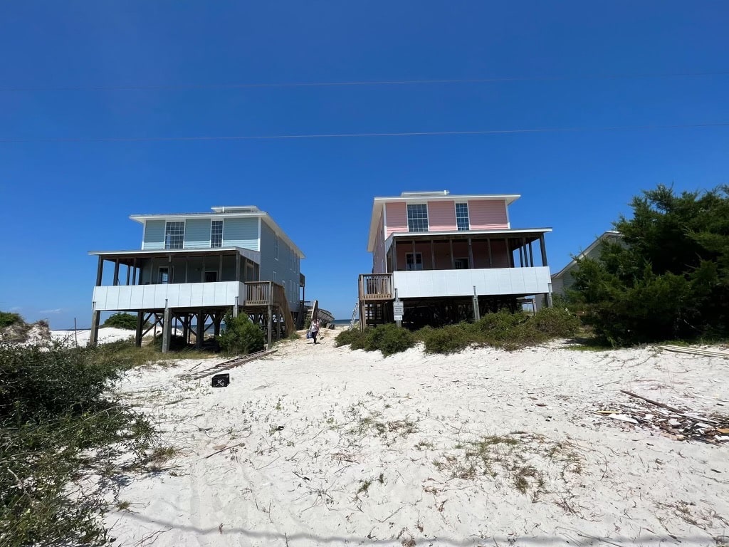 Two beachfront houses on stilts sit side by side on a sandy beach under a clear blue sky. The left house, with its roofing, is painted blue, while the right house is pink. A wooden walkway leads from the beach towards the houses, and dense greenery is seen on the right.