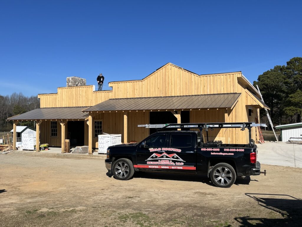 A wooden building under construction with a person working on the roofing. A black truck with the Halo Roofing logo and contact information is parked in front. Construction materials are scattered around the area, and the sky is clear and blue.