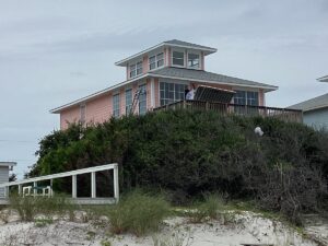 A two-story pink house with large windows sits atop a sandy hill covered in dense shrubbery. A person is on the deck near the house. The sky is overcast, giving the scene a greyish tone. A white handrail leads up the slope, and beach grass is visible in the foreground, accentuating the house's roofing.