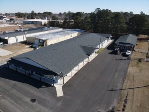 Aerial view of a large, single-story building with a dark grey roof, possibly updated by Halo Roofing, and multiple entrances. The structure is surrounded by a paved area, with a smaller adjacent building on the right. Trees and additional industrial buildings are visible in the background.