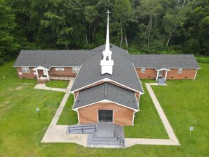 Aerial view of a small brick church with a white steeple, surrounded by green lawn and trees. The church has three adjoining sections and multiple entrances, with a concrete path leading to the main door. Roofing is visible on the structure. Handicapped parking signs are near the entrance.