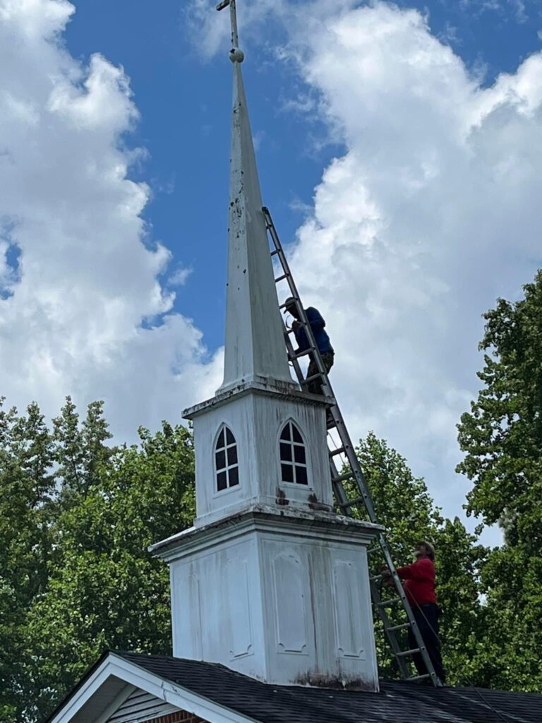 Two workers on ladders are performing maintenance on the white steeple of a building resembling a church, possibly for Halo Roofing. The sky is partly cloudy, and there are green trees in the background.