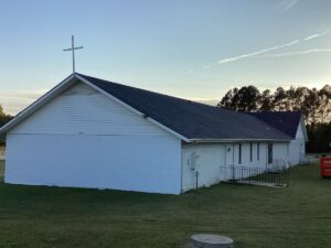 A white, single-story church with a dark roofing and a cross mounted on the peak. The building is set against a backdrop of trees and grass, with a clear sky in the background. A ramp leads to an entrance on the side.