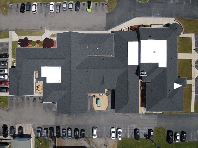 Aerial view of a large building with gray roofing and white sections, surrounded by parked cars and landscaped areas. The building appears to be part of a larger complex with multiple entrances and pathways. The parking lots have numerous vehicles.