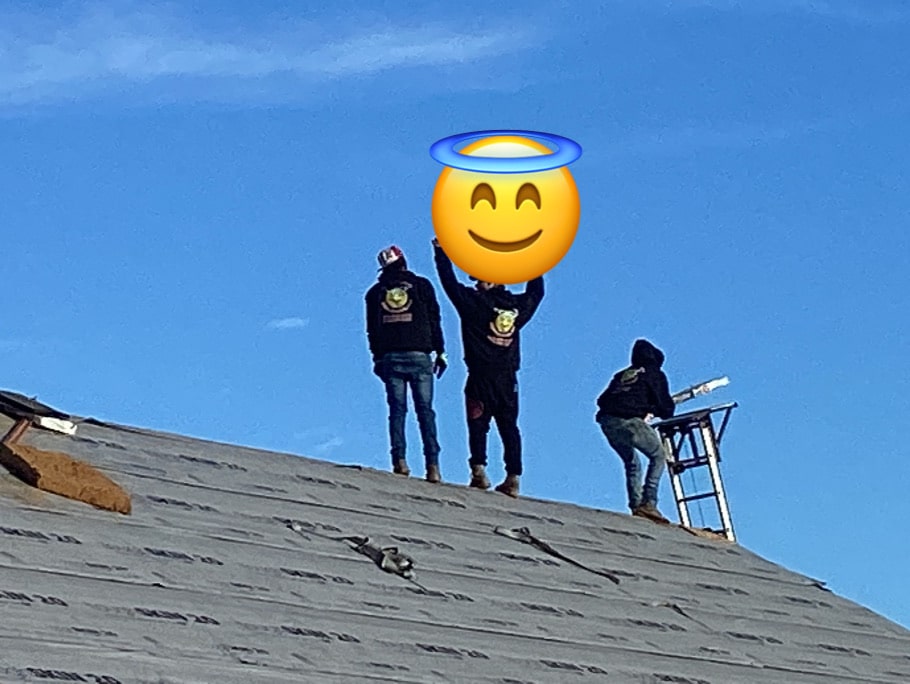 Three workers are on a roof under a clear blue sky. Two are standing, and one is kneeling near a ladder, all wearing protective clothing. The face of the standing worker on the left is covered with an emoji of a smiling face with a halo, fitting for this halo roofing team.