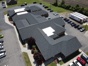 Aerial view of a building complex with dark roofing and light pavement surrounding it. Several cars are parked around the complex, and a large truck with the logo "Sysco" is visible on the right. The landscape includes grass, trees, and a field in the background.