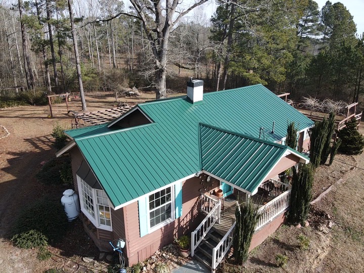 Aerial view of a single-story house with a green metal halo roofing, surrounded by trees. The house has a small front porch and a chimney. The yard features shrubs, a pergola, and is bordered by a forest. The overall scene shows a wooded and serene environment.