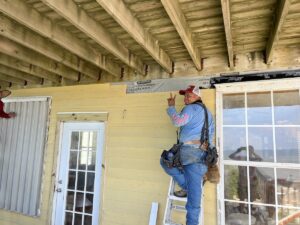 A construction worker wearing a blue shirt, jeans, and a tool belt stands on a ladder, working on the ceiling of a yellow house. He is near a glass door and next to a window. The exterior features siding and wooden beams above, indicative of careful roofing craftsmanship by Halo Roofing.