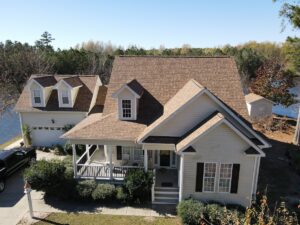 A two-story house with beige siding and a brown shingled roof, expertly installed by Halo Roofing. It has a covered front porch, two dormer windows, and an attached garage with two doors. The property is surrounded by trees and is adjacent to a body of water. A black car is parked in the driveway.