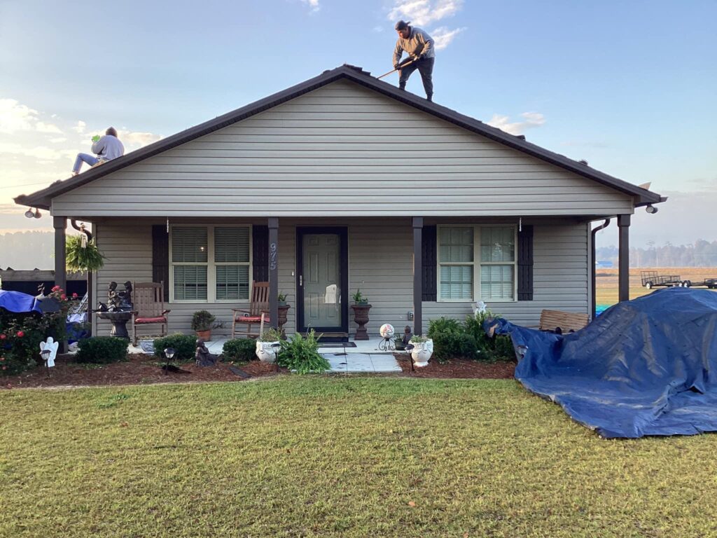 Two people work on the roof of a beige single-story house by Halo Roofing. The porch has two chairs with small tables, and the front yard boasts potted plants and garden decorations. A large blue tarp covers sections of the lawn. The sky is clear and bright in the background.