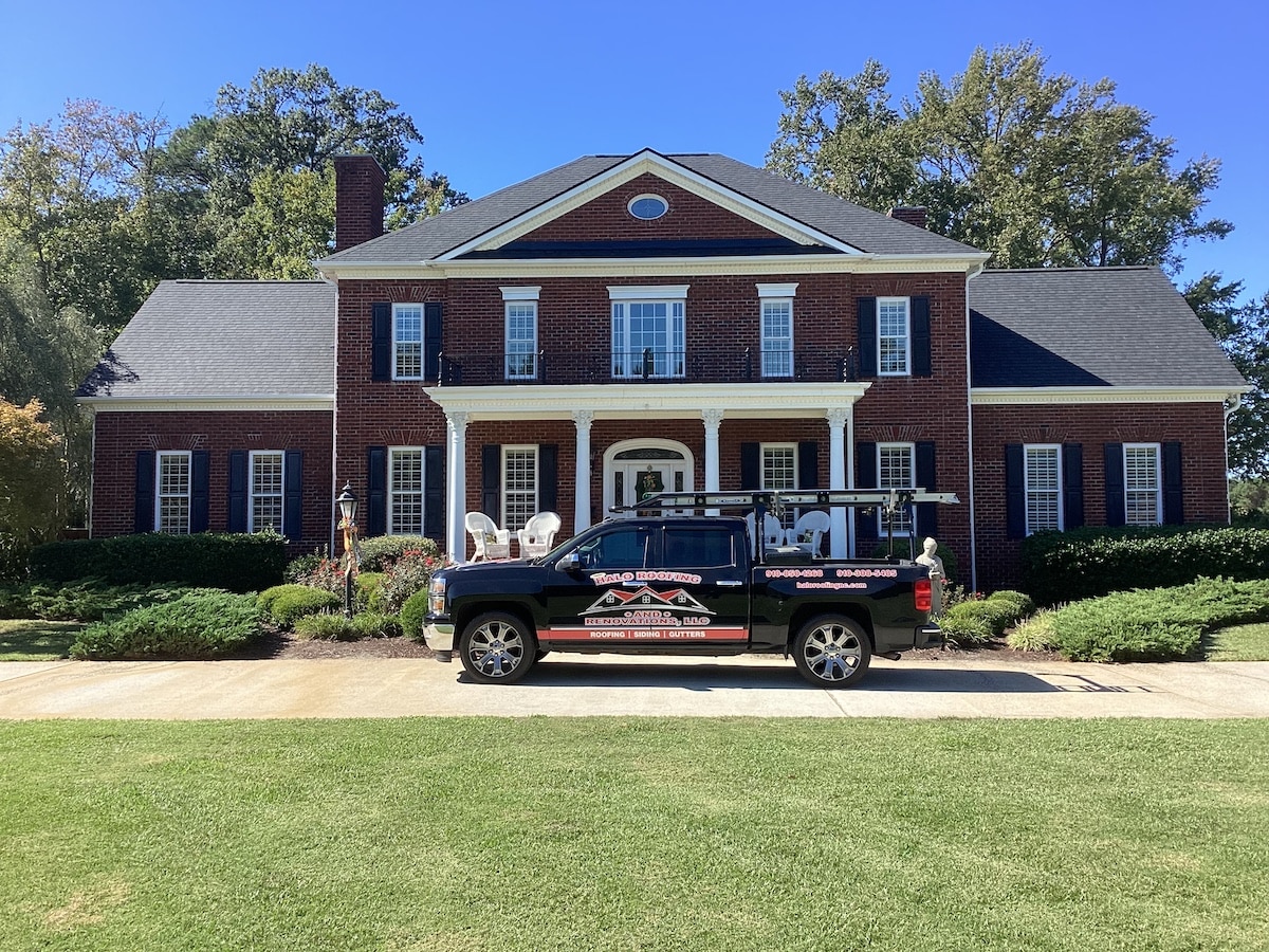 A two-story brick house with white accents and large windows is shown. A black utility truck with ladders and equipment on the roof, featuring Halo Roofing branding on the side, is parked in the driveway. The front yard is landscaped with bushes and a well-manicured lawn.