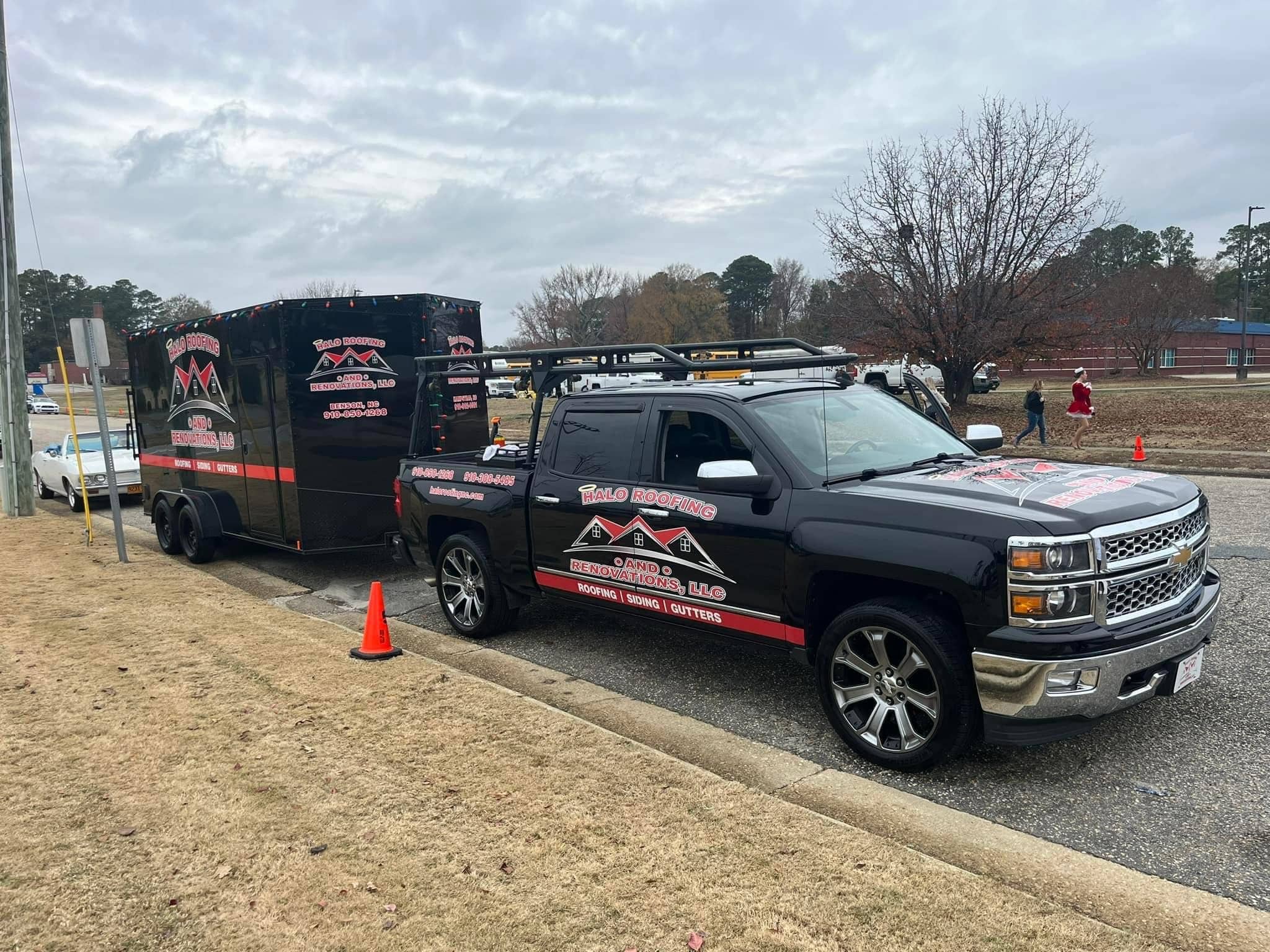 A black pickup truck with a matching enclosed trailer, both branded with "ROAD ROGUE EXTERIORS, LLC" and contact information, is parked on the side of a road near an orange cone. Trees and people walking are visible in the background, hinting at another successful Halo Roofing project in progress.