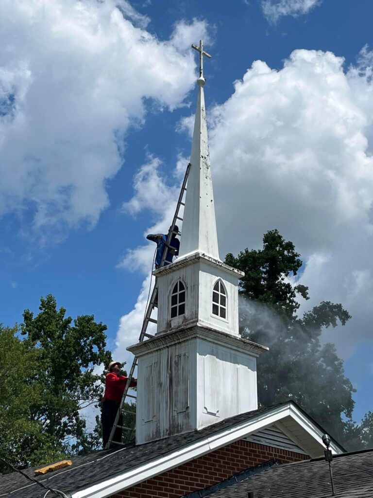 Two people work on the steeple of a church. One person in a red shirt stands on the roof and uses a tool, showcasing their roofing skills, while the other person in a blue shirt stands higher up on a ladder. The steeple is white with windows, and smoke or dust is visible. The sky is partly cloudy.