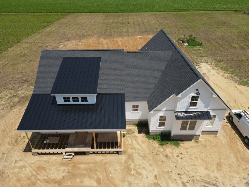 Aerial view of a newly constructed white house with a black roof by Halo Roofing, situated on a dirt lot. The house features multiple gables and partially completed wooden decking. A white vehicle is parked next to the house, and the surrounding area includes a grassy field and farmland.