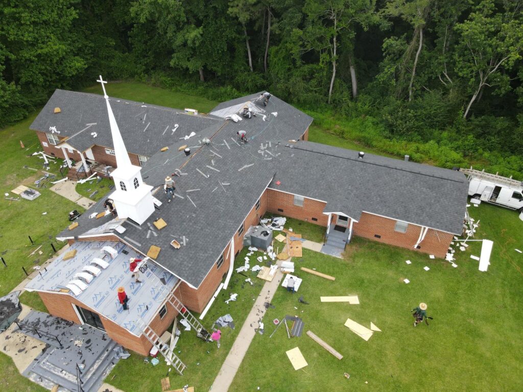 A bird’s-eye view of a church with a damaged roof under the expert care of halo roofing workers. Several workers are on the roof, diligently repairing it. Debris and construction materials are scattered around the building. The surrounding area is lush with trees in the background. Ladders and tools are visible.