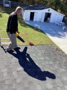 A man wearing a black sweater and khaki pants uses a leaf blower to clear leaves off a rooftop. His shadow is visible on the roof of the Halo Roofing project. Below, a well-kept lawn and a white shed with open doors can be seen, with trees in the background.