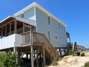 A beach house with light blue siding, elevated on wooden stilts with a wooden staircase leading up to a covered porch. Sand dunes and sparse vegetation surround the house, and the clear blue sky is visible above, highlighting the expertly crafted halo roofing. The ocean can be seen in the background.