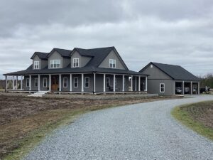 A modern gray house with a front porch and an adjacent detached garage sits on a gravel driveway. The house features roofing with a gable roof and dormer windows. The surrounding area appears to be a rural setting with an overcast sky.