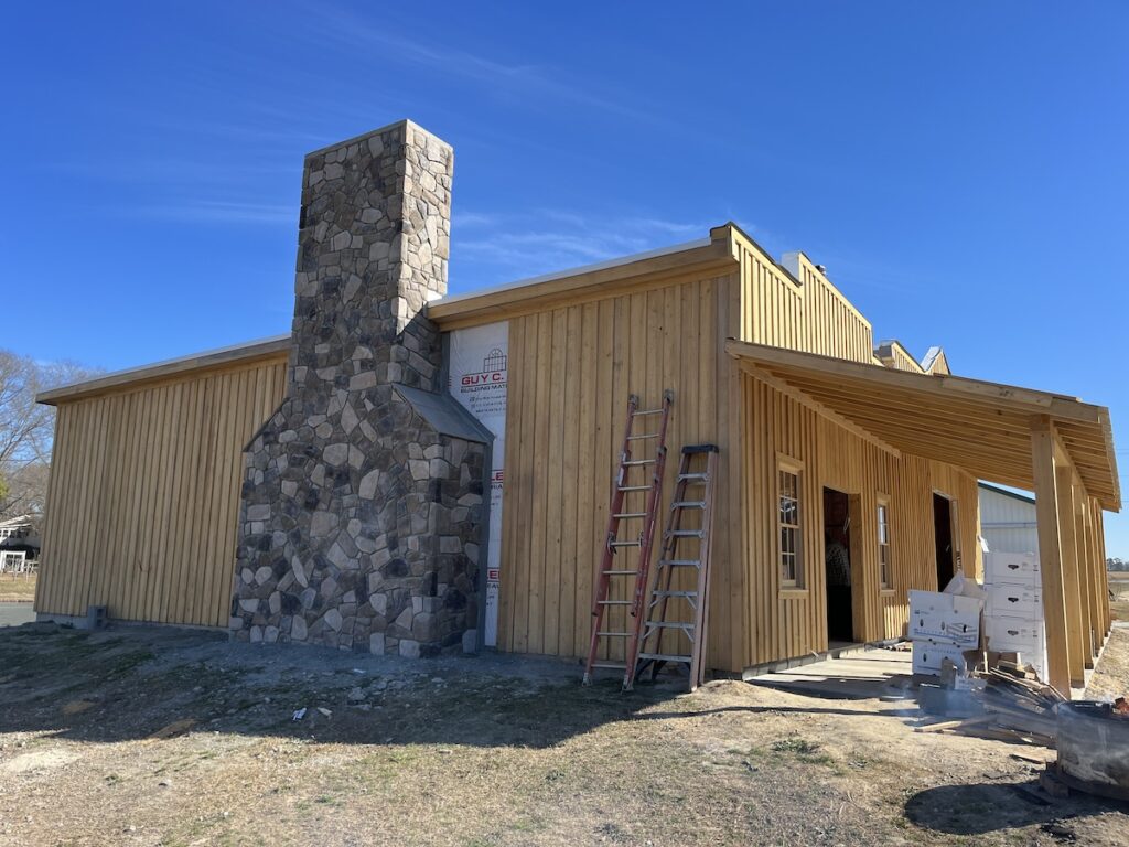 A wooden building with vertical siding is under construction. It has a large stone chimney on one side and halo roofing being installed. There's a ladder leaning against the building, stacks of construction materials, and a partially covered porch. The sky is clear and blue.