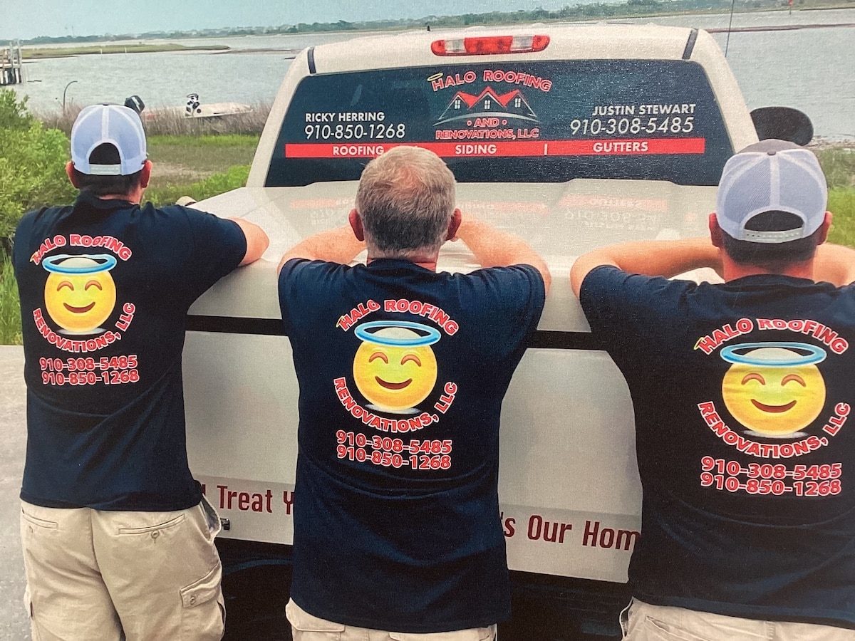 Three people in matching "Halo Roofing Renovations, LLC" shirts lean on the back of a pickup truck. The truck features the company's logo, contact information, and roofing services offered. A body of water and grassy area are visible in the background.