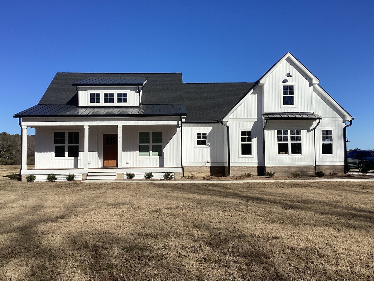 A newly-constructed modern farmhouse-style home with white exterior walls, roofing in sleek black, and a wooden front door. It features a front porch with steps, multiple windows, and a manicured lawn under a clear blue sky.