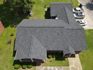 Aerial view of a residential home with a dark gray shingled roof by Halo Roofing. The house has a porch and is surrounded by a well-kept lawn with trees and shrubs. Several vehicles are parked in the driveway and on the street. The weather is clear and sunny.