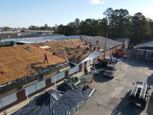 A view of a building with workers on the roof conducting repairs for Halo Roofing. Some areas of the roof are stripped down to the wooden deck, while others are partially covered with black roofing material. Tarps, ladders, and equipment are visible surrounding the building.