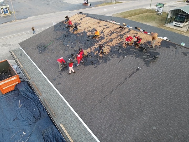 A group of workers from Halo Roofing is repairing the roof of a building. They are removing old shingles and preparing the roof for new ones. Some of them are wearing red shirts. There is a dumpster on the ground filled with debris, and the area around the building is clear.