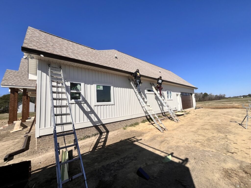 Two workers stand on ladders leaned against the side of a white building under construction. The building, which is getting new halo roofing, has a pitched roof and multiple ladders are positioned around it. The ground around is bare dirt, and the sky is clear and blue.