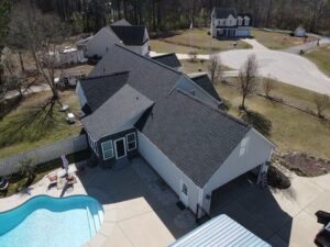 Aerial view of a suburban house with a large, fenced-in backyard featuring a swimming pool surrounded by a patio area. The roofing is in excellent condition. Nearby, there are other houses and a wooded area in the background.