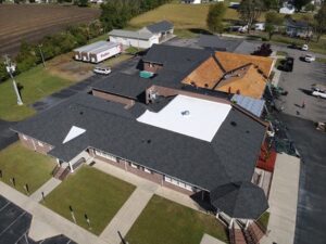 Aerial view of a large building with grey shingles and a white section of the roof. Part of the roof, managed by Halo Roofing, is under construction, with workers and materials visible. Surrounding the building are parking lots, a grassy field, and other nearby structures.